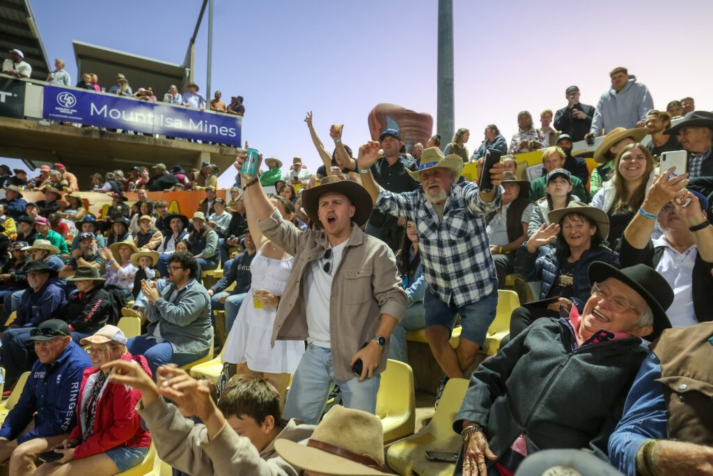 A large crowd of people, many wearing hats, cheer enthusiastically while seated in stadium stands during an outdoor event.
