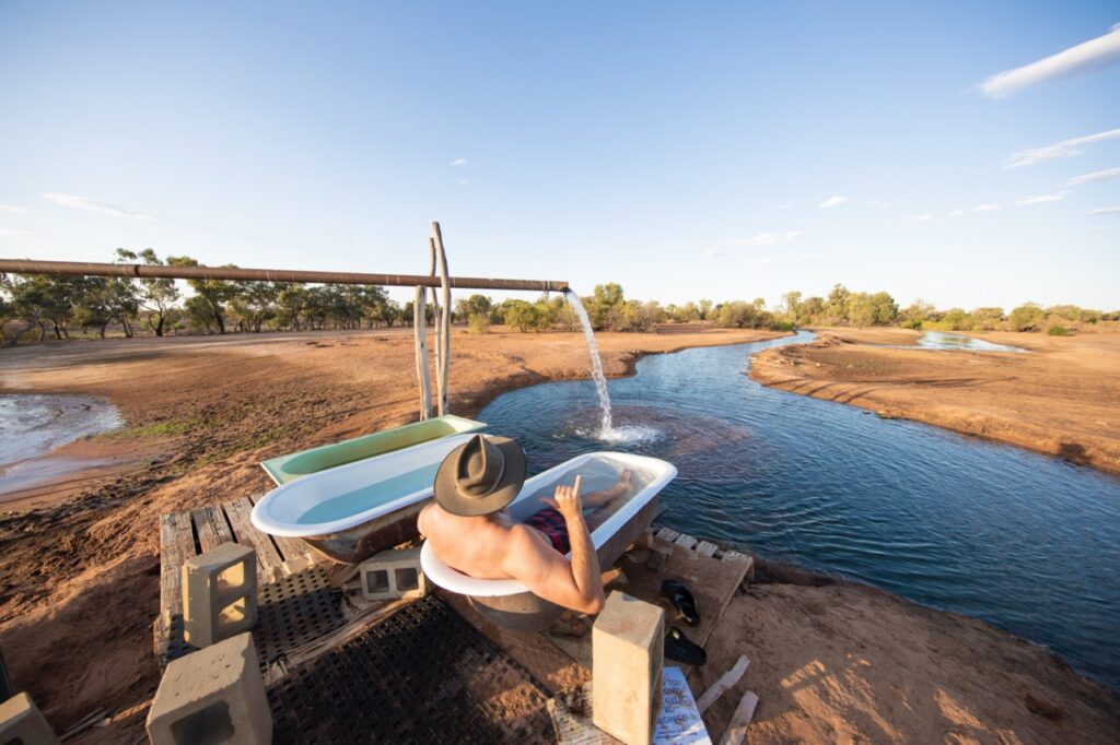 Person in a hat relaxes in an outdoor bathtub filled with water from a pipe, set beside a river and surrounded by a dry, open landscape.