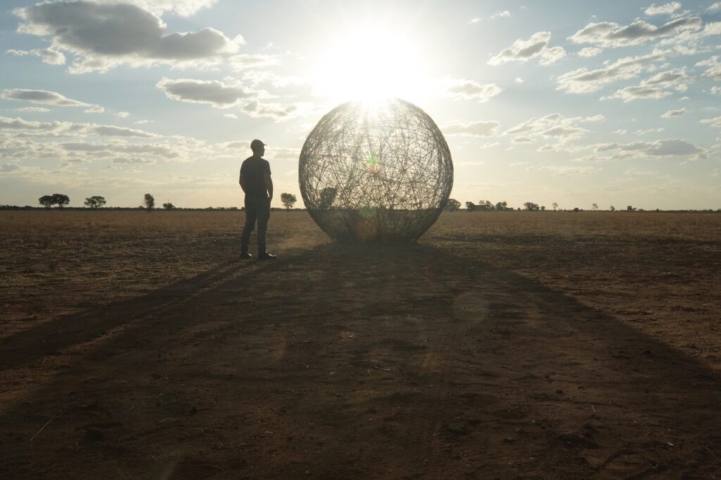 A person stands in front of a large, spherical wire sculpture in a vast, open field at sunset.