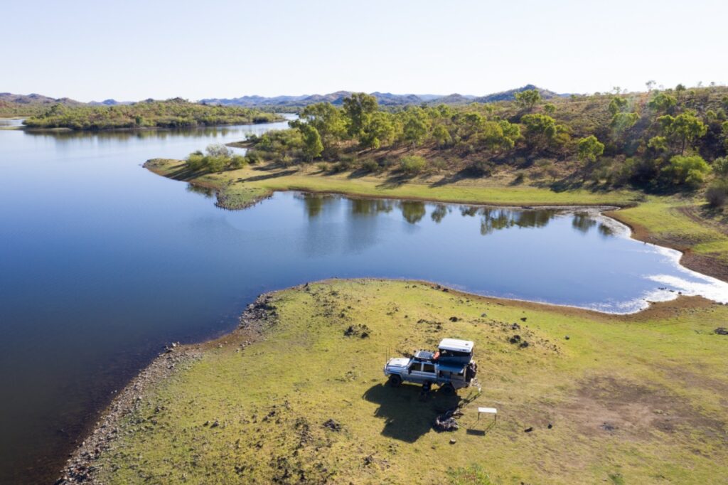 Aerial view of a lake with a parked SUV and trailer on grassy shore, surrounded by hilly terrain and sparse trees. Clear sky above.