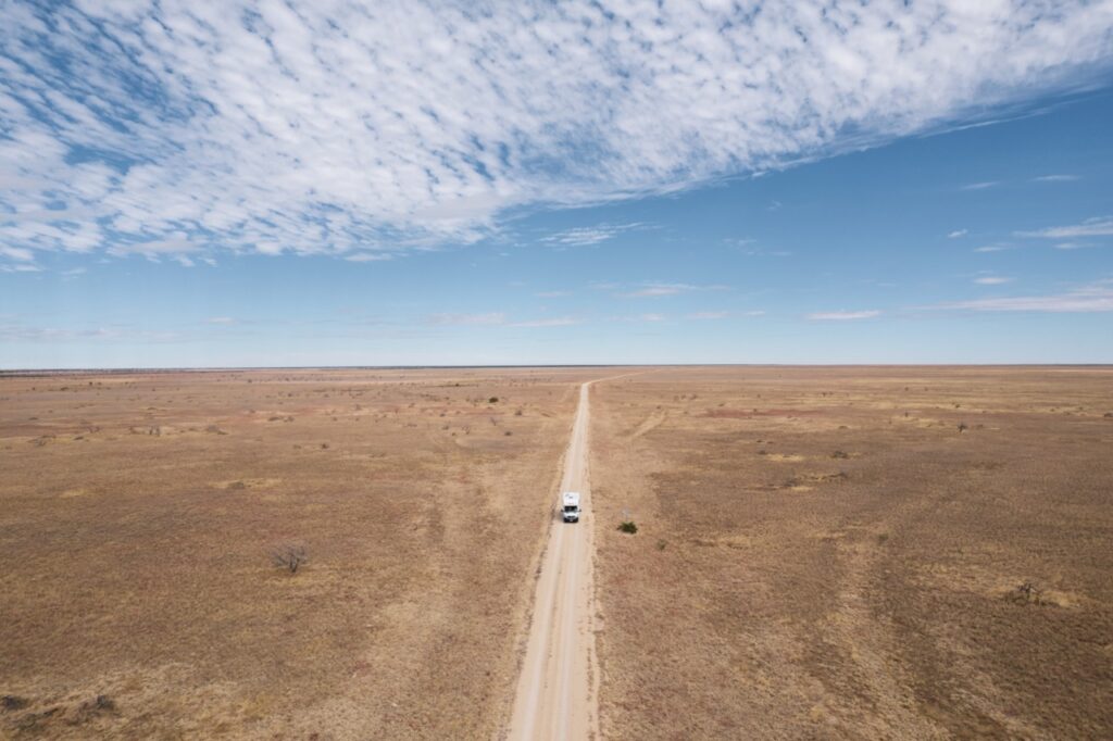 A white vehicle travels on a narrow dirt road through an expansive, flat, and dry landscape under a partly cloudy sky.