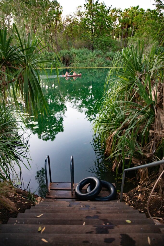 Wooden steps lead down to a serene pond surrounded by lush greenery, with an inflatable tube placed on the steps and two individuals canoeing in the water.