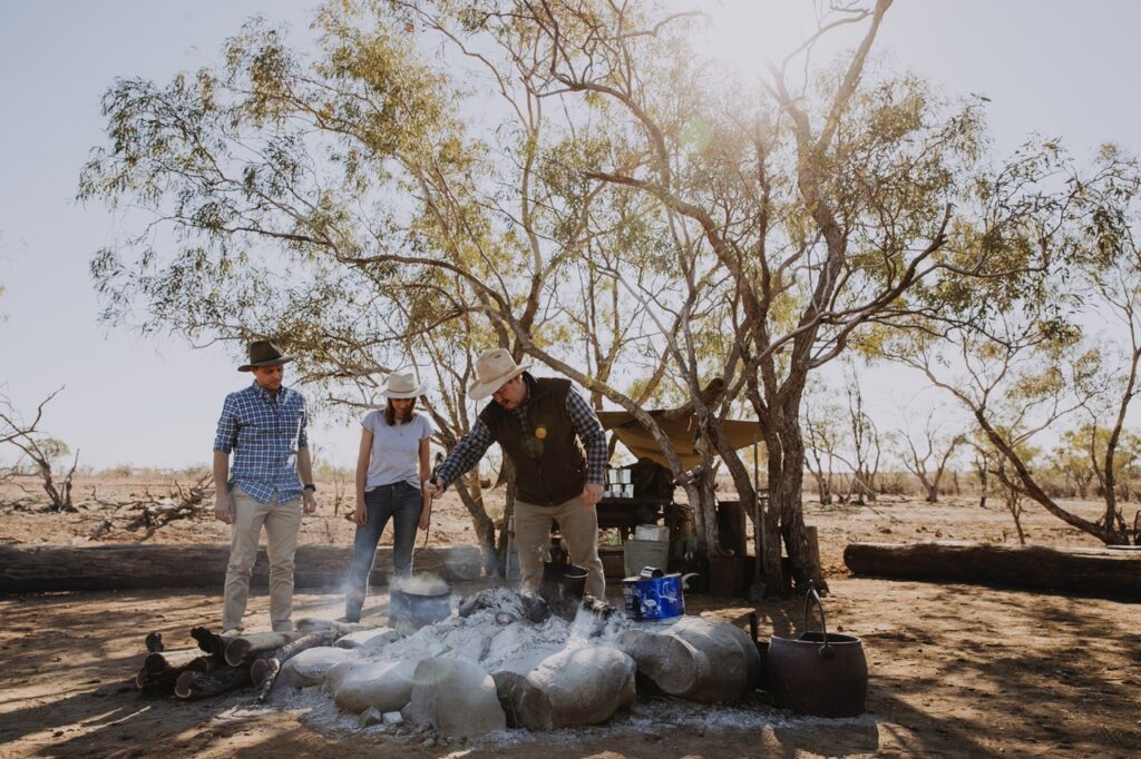 Three people wearing hats are standing around a cooking area in an outdoor woodland setting. They appear to be cooking food over a fire pit made of stones. Trees and camping gear are in the background.