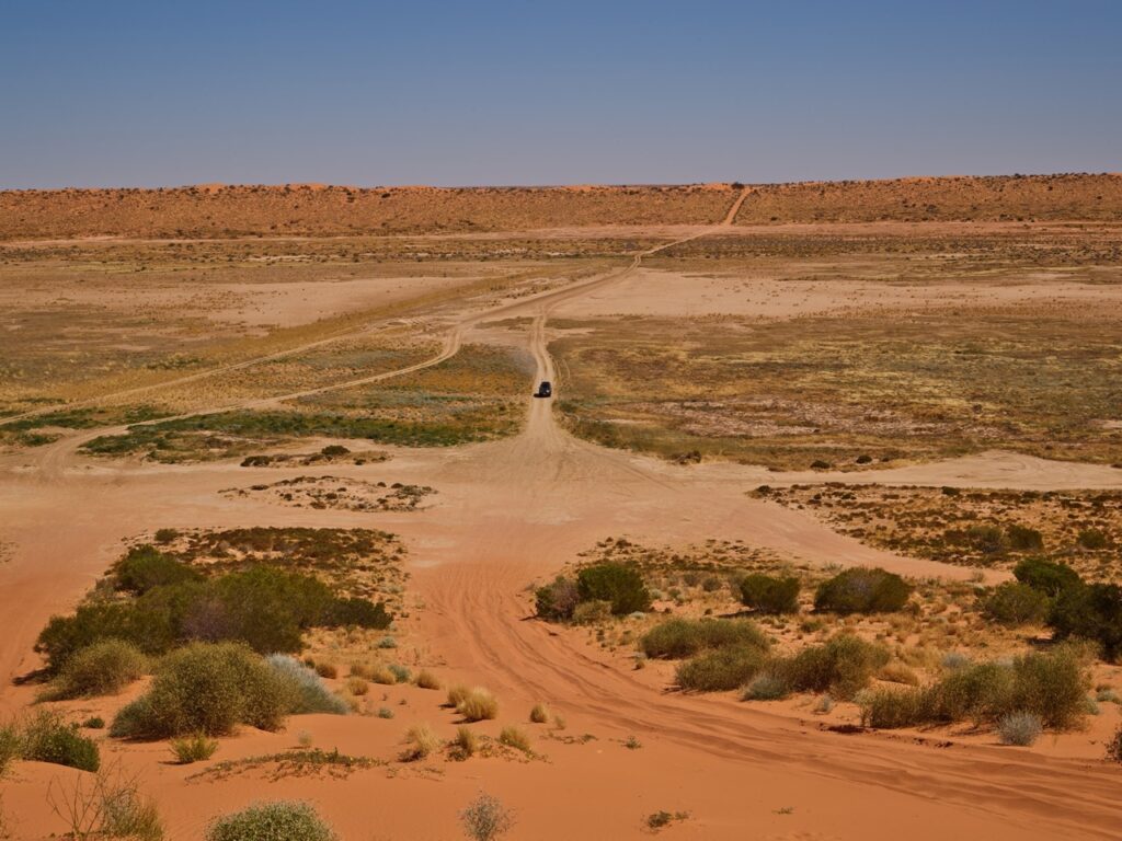 A lone vehicle drives along a dirt road in a vast, arid desert landscape with sparse vegetation, under a clear blue sky.