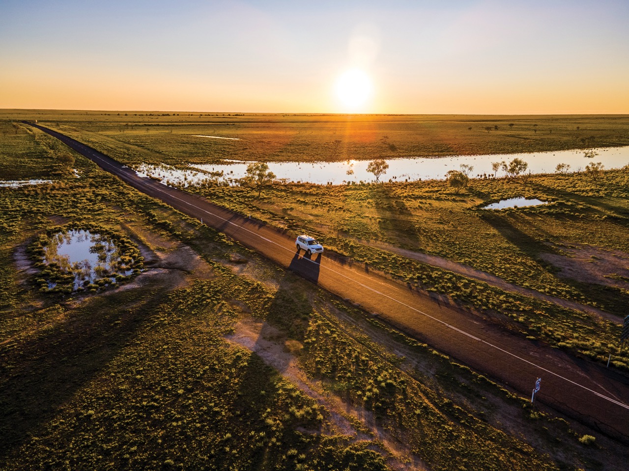 A white vehicle travels on a road through a rural landscape at sunset, with fields, a large puddle, and sparse trees visible in the background.