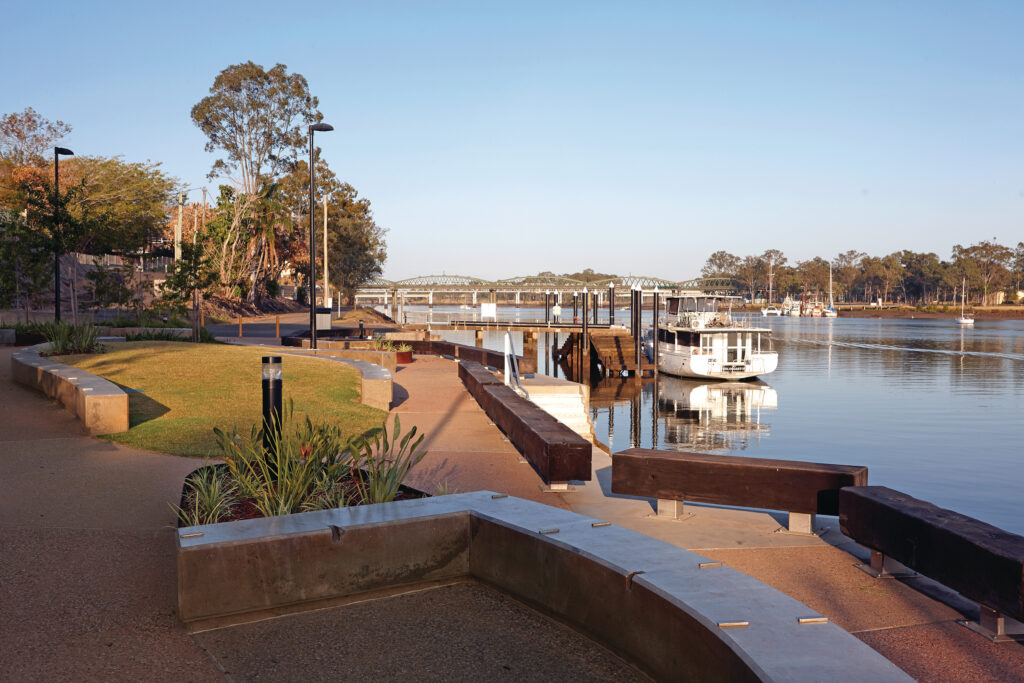 A riverside scene featuring a paved walkway, benches, greenery, and a docked white boat. A calm river and a distant bridge are visible under a clear blue sky.