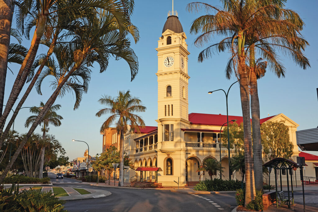 A street view of a historic building with a clock tower surrounded by palm trees, illuminated by warm sunlight.