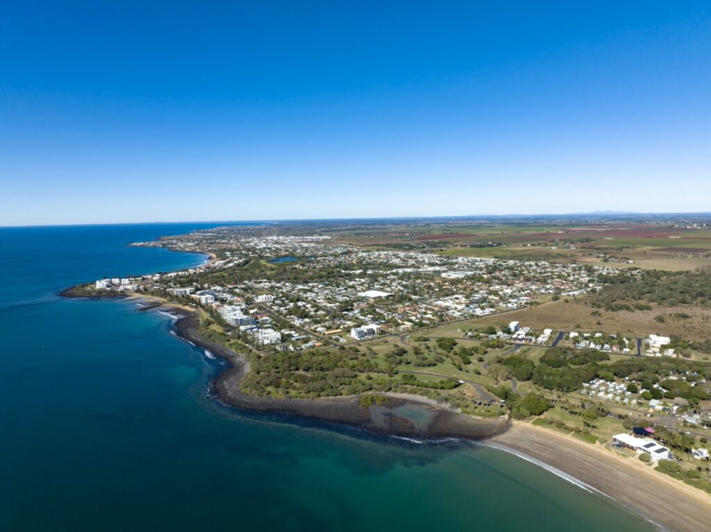 Aerial view of a coastal town with a sandy beach, turquoise sea, residential areas, green spaces, and farmlands extending into the horizon under a clear blue sky.