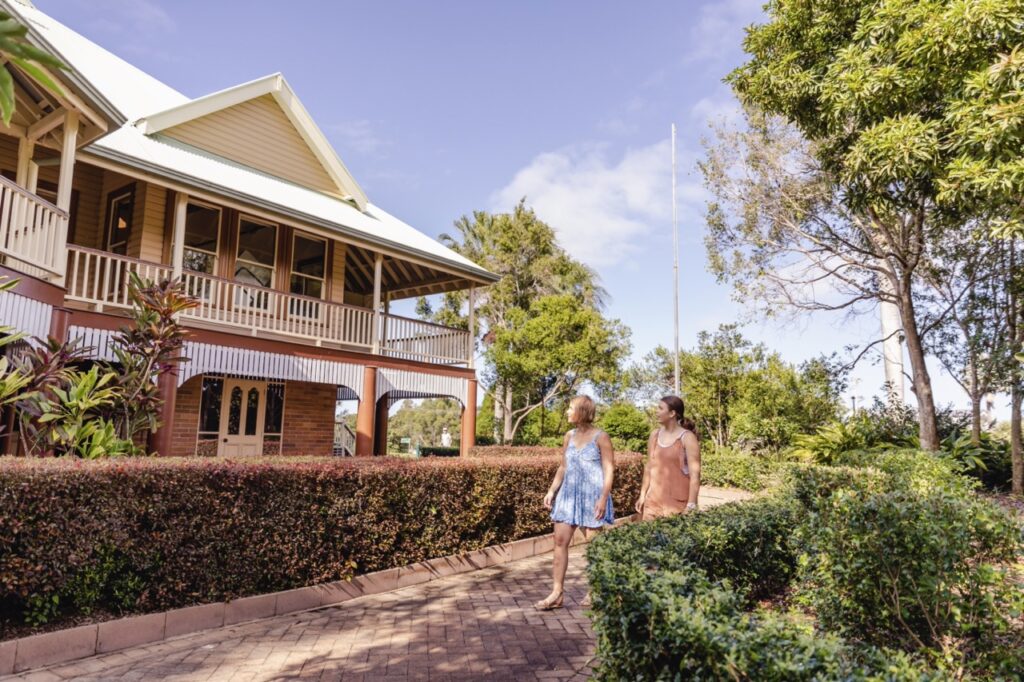 Two people walk along a path next to a large, wooden house with a wraparound porch, surrounded by lush greenery on a sunny day.