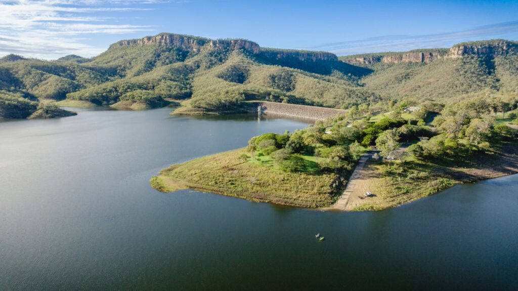 Aerial view of a dam surrounded by water, dense green vegetation, and mountainous terrain under a clear blue sky.