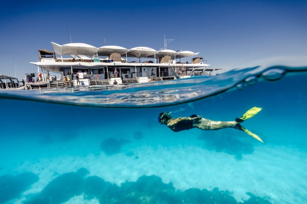 A snorkeler swims underwater near a floating dock structure on a clear, sunny day with several people on the platform above the water.