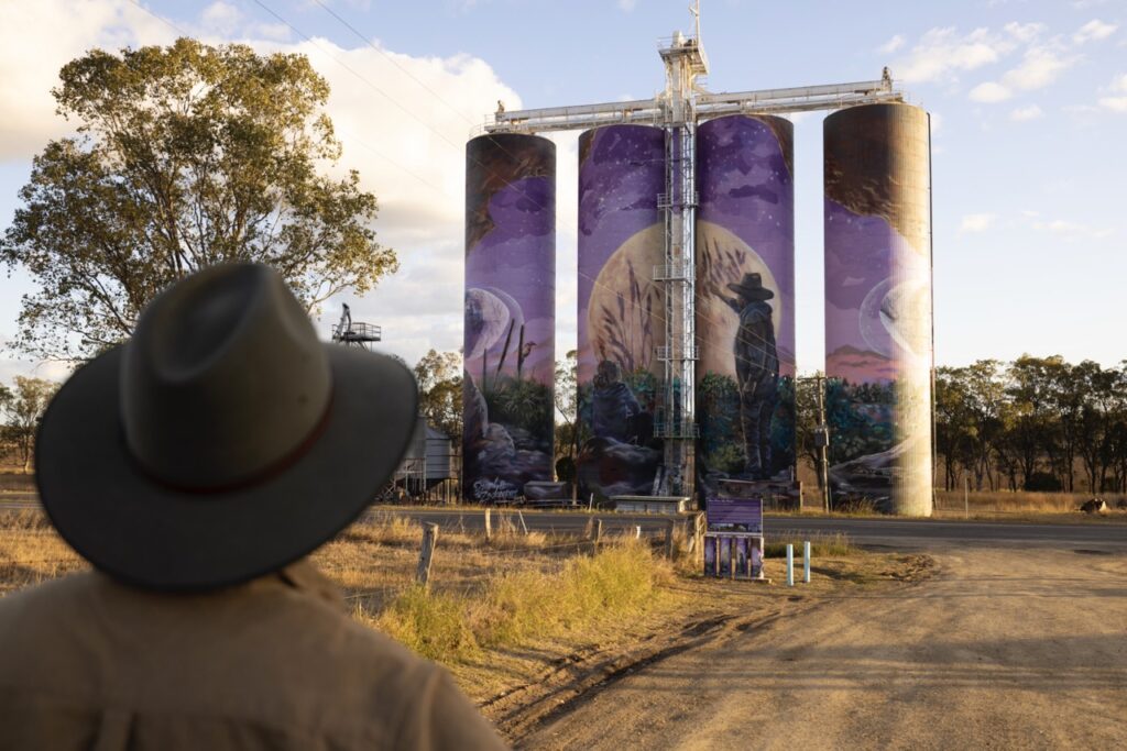 A person in a hat looks at grain silos with a large mural depicting a cowboy and nature scene.