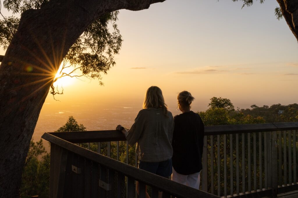 Two people stand on a wooden lookout platform at sunset, facing a scenic view with a sunburst peeking through tree branches.