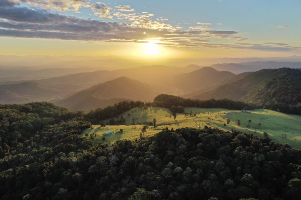 Aerial view of a green valley and rolling hills at sunrise with scattered trees and distant mountains under a partly cloudy sky.