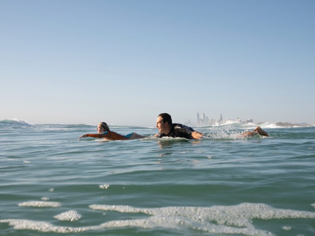 Two people paddle on surfboards in the ocean, with a distant city skyline visible on the horizon under a clear blue sky.