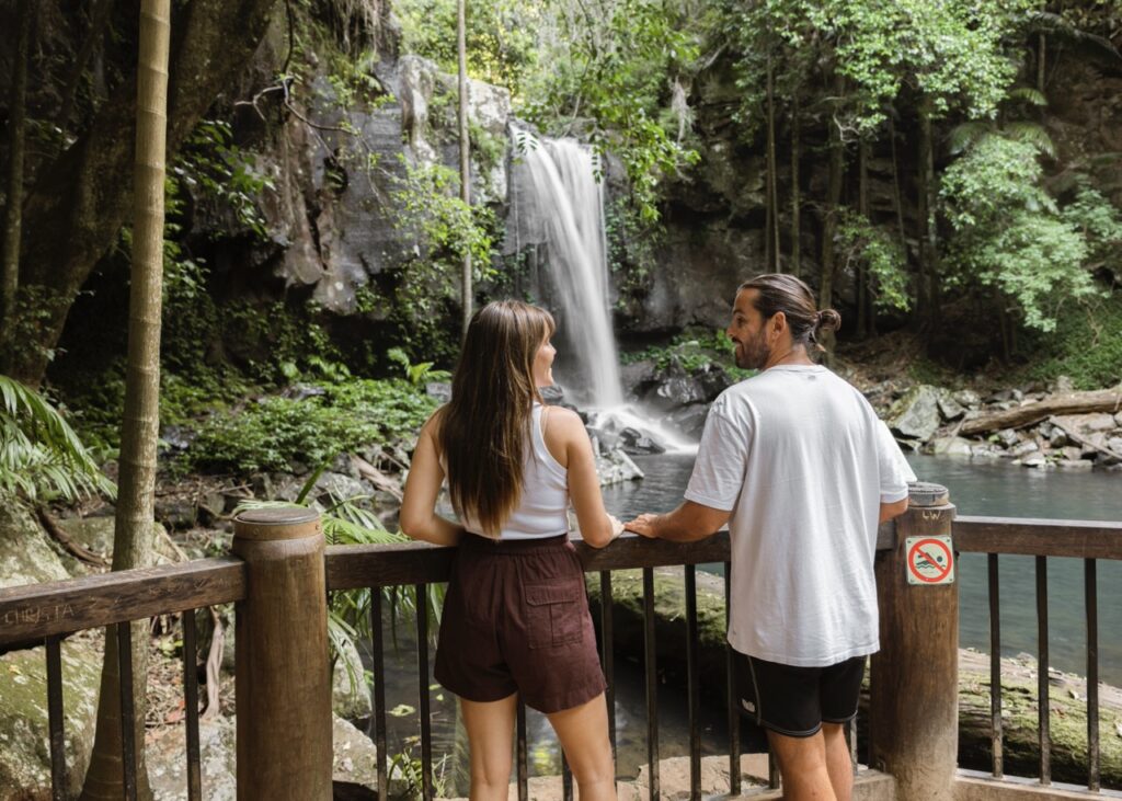 Two people stand on a wooden platform, looking at a waterfall surrounded by lush greenery in a tropical forest.