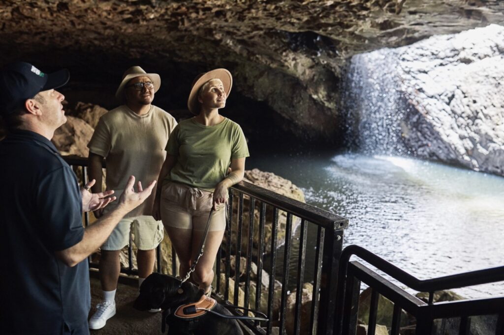 Three people, one with a guide dog, stand near a railing inside a cave, looking at a waterfall. A guide appears to explain something to them.