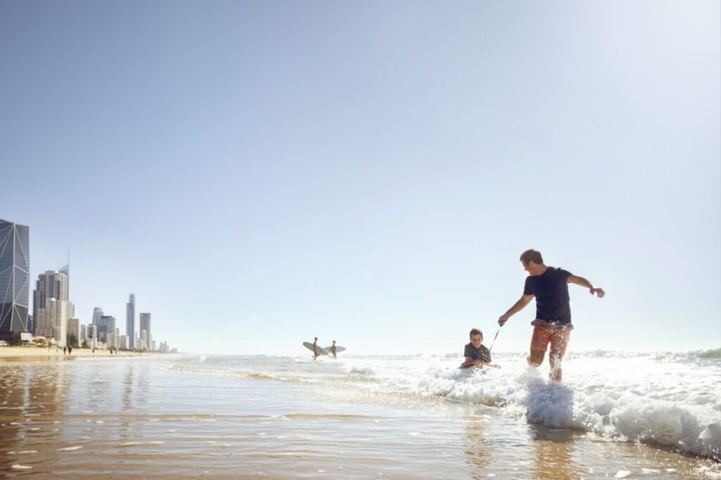 Two people play in shallow ocean water at a beach on a sunny day. Skyscrapers and buildings line the shore in the background, while other people with surfboards enter the water.