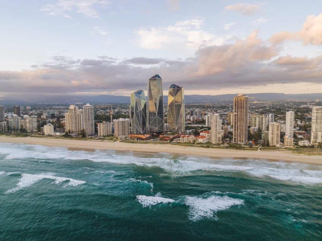 Aerial view of a coastal city with tall skyscrapers near the beach, waves breaking on the shore, and a partly cloudy sky overhead.