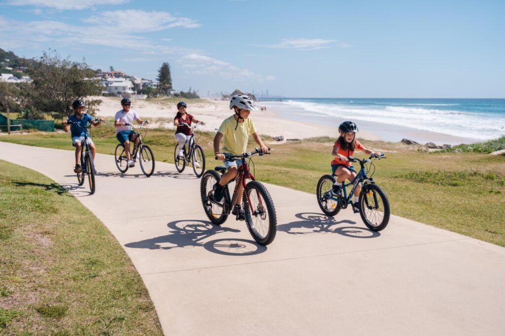 A group of people, including children and adults, ride bicycles on a seaside path under a clear blue sky.