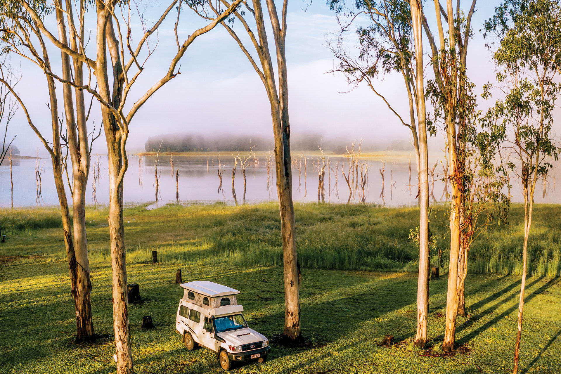 An off-road vehicle parked on a grassy area near a lake bordered by tall trees. Fog covers the distant background.