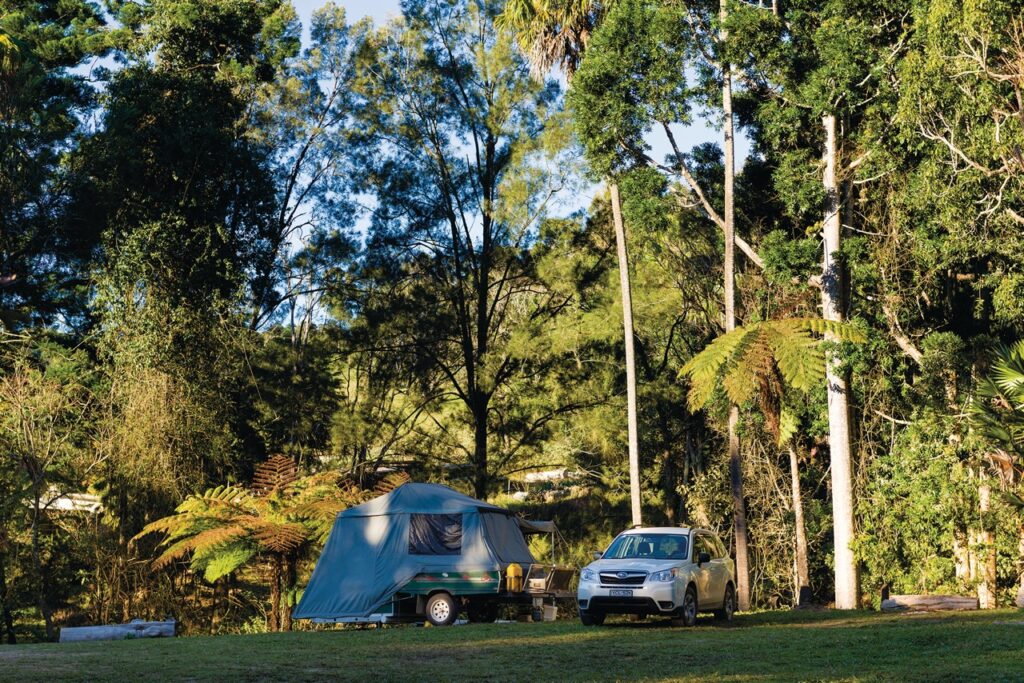 A small green tent trailer is set up among tall trees in a forested area, with a white SUV parked beside it.