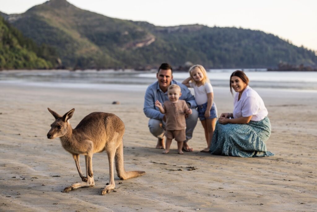 A family with two young children kneels and waves to a kangaroo on a beach, with wooded hills in the background.