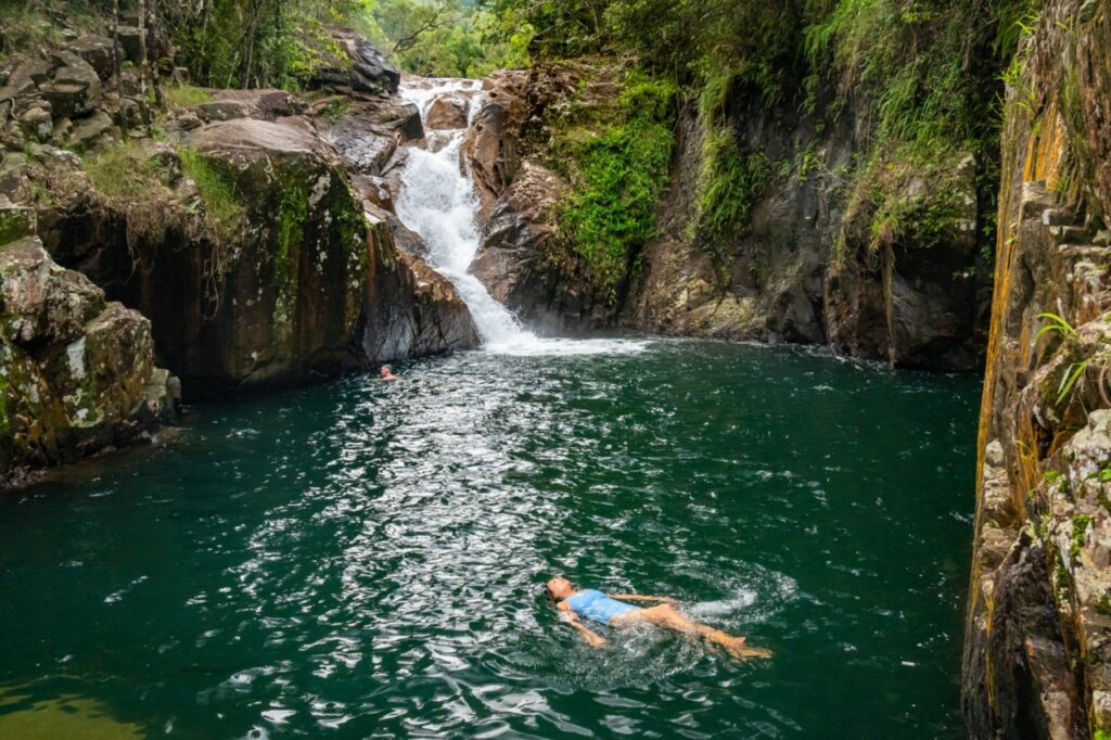 A person in a blue swimsuit is swimming in a natural pool with a small waterfall, surrounded by rocks and lush greenery. Another person is visible in the water near the waterfall.