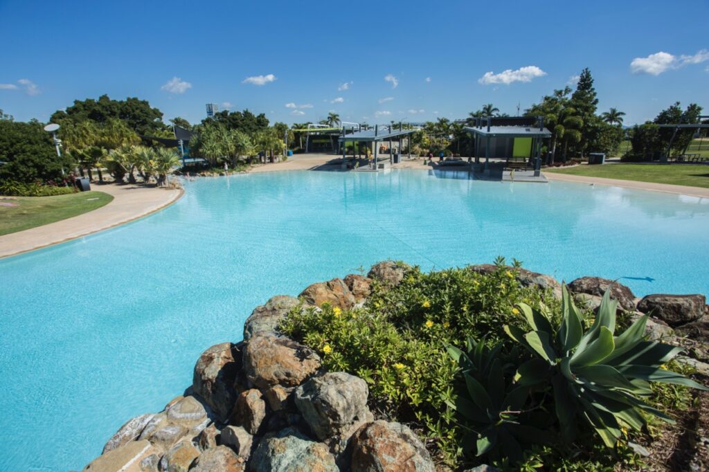 Large outdoor swimming pool with clear blue water, surrounded by greenery, rocky landscaping, and modern shaded seating areas under a clear blue sky.