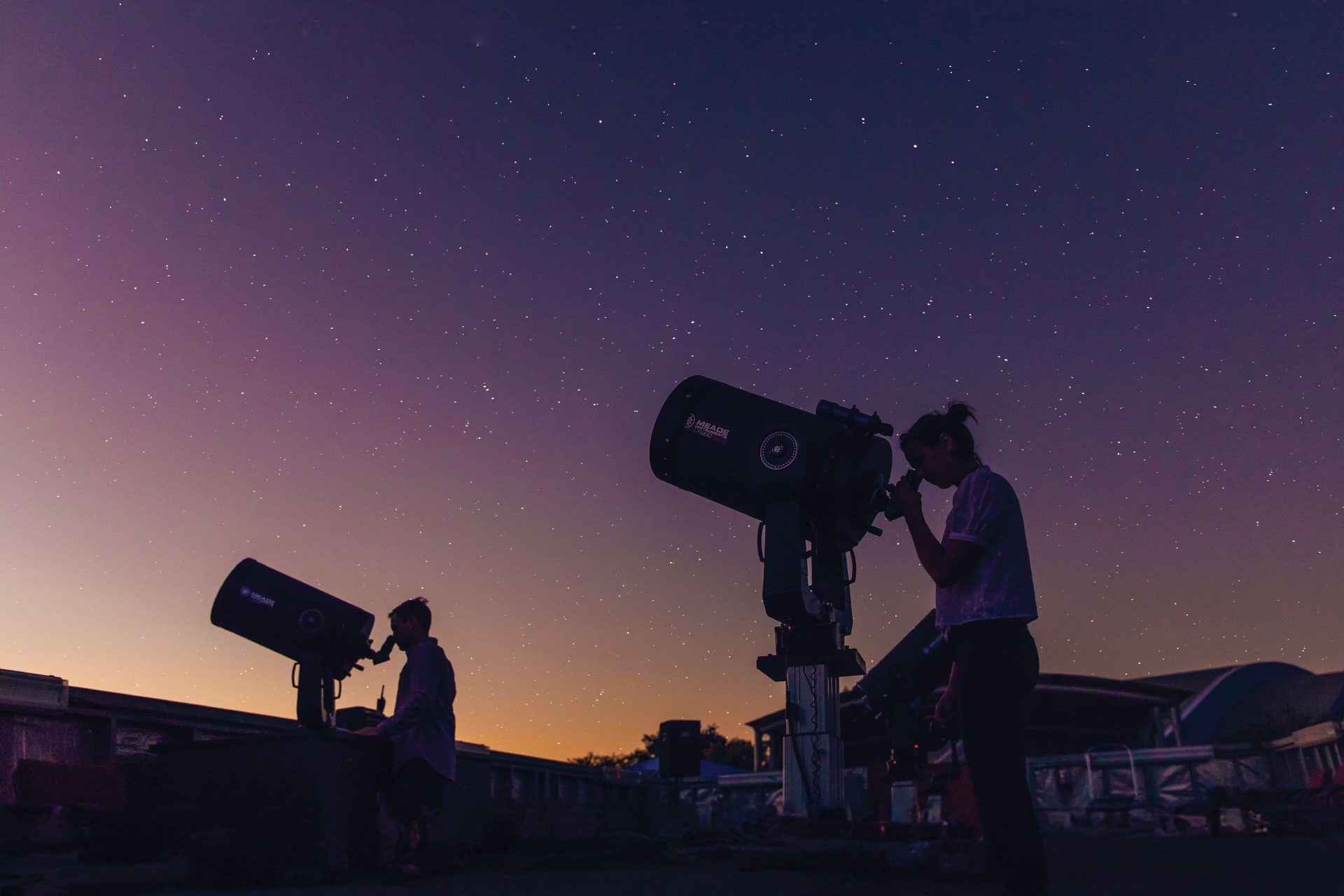 Two silhouetted individuals using telescopes to observe the starry night sky against a twilight backdrop.