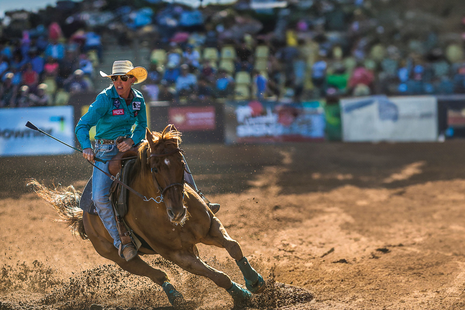 A rodeo rider in a teal shirt maneuvers a horse through a dusty arena during a competition, with spectators in the background.