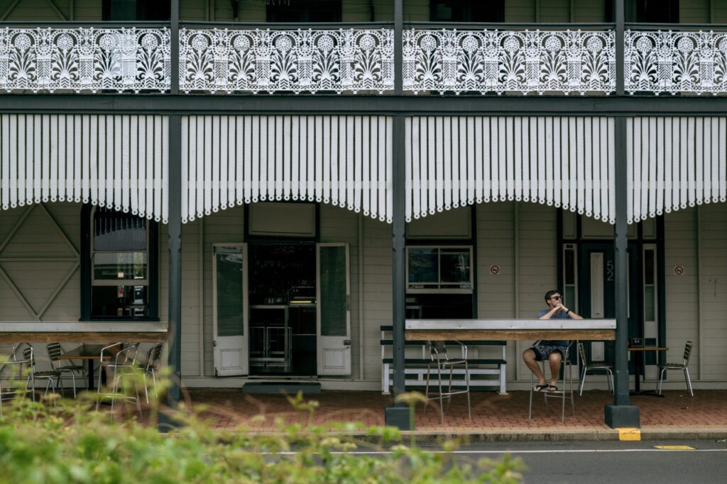 A person sits alone at an outdoor table under a decorated balcony and striped awning on a building with metal tables and chairs. The sidewalk is visible in the foreground.