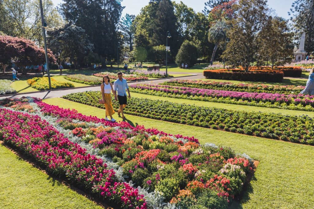 A couple strolls through a well-maintained garden with vibrant flower beds and green lawns on a sunny day.