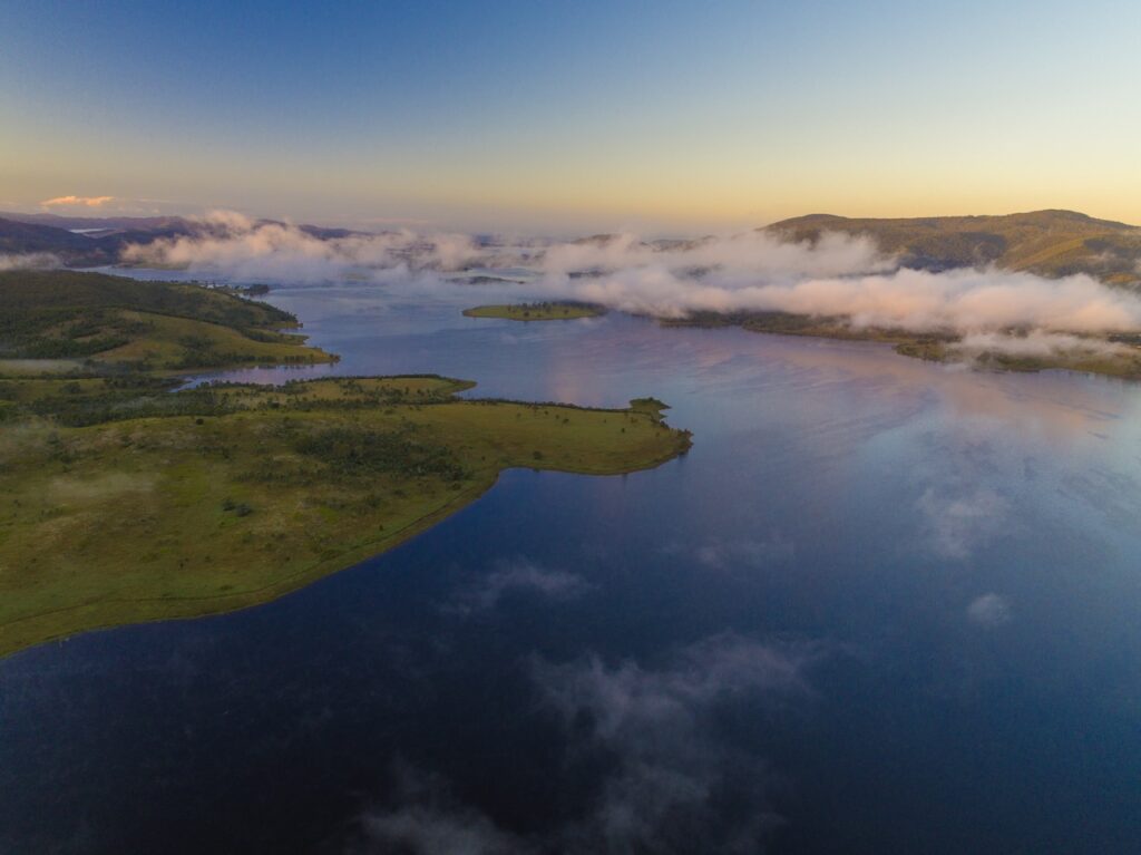 Aerial view of a serene lake surrounded by hilly terrain and greenery, with patches of low-lying clouds near the water and a clear sky at dawn.