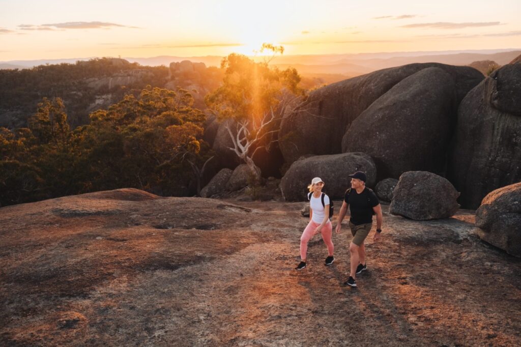 Two people hiking on rocky terrain during sunset, with trees and distant hills in the background.