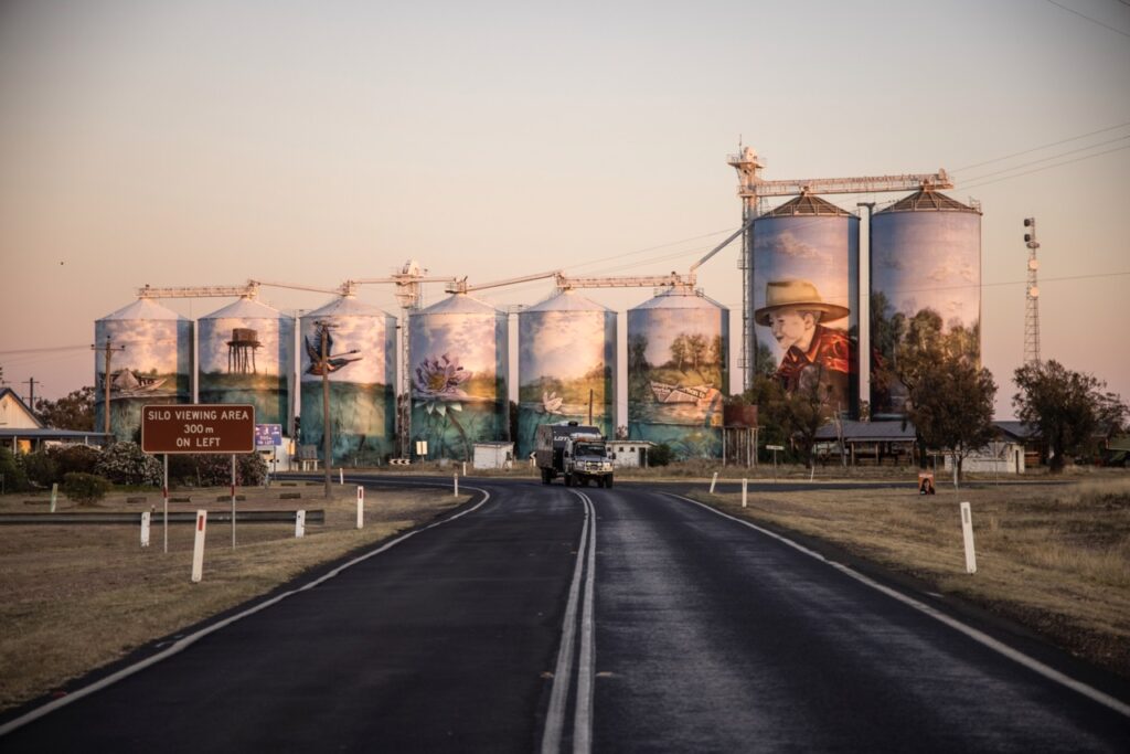 A row of silos painted with murals, including a portrait, birds, and a sunset scene, viewed from a nearby road with a sign indicating a silo viewing area 300 meters to the left.