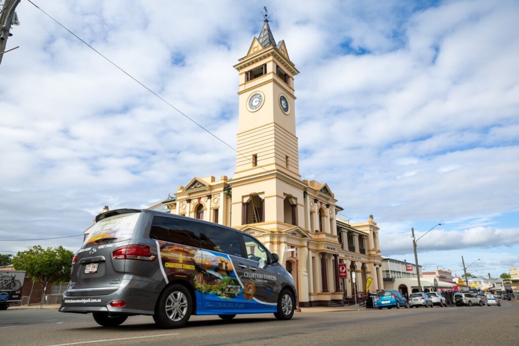 A van drives on a road in front of a historic building with a clock tower under a partly cloudy sky.