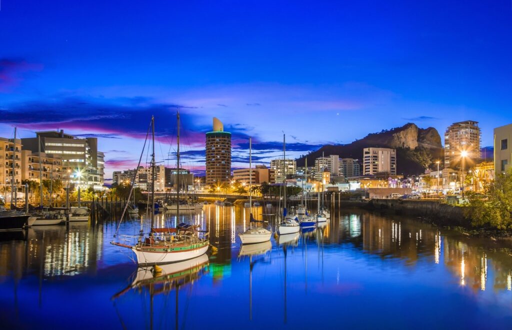 A marina at dusk with sailboats docked in calm water; city buildings and a rocky hill are illuminated in the background.