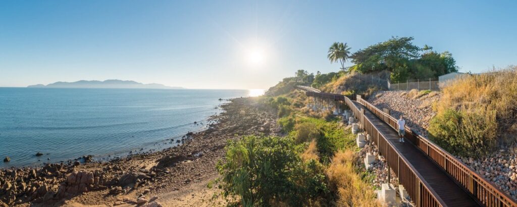 A coastal boardwalk stretches alongside rocky shoreline and sparse vegetation, with a person walking and the sun low in the sky over calm ocean waters. Hills are visible in the distance.