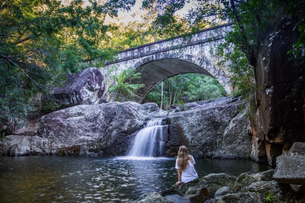 A person with long hair sits on rocks near a small waterfall and pond, with a stone bridge spanning overhead in a forested area.