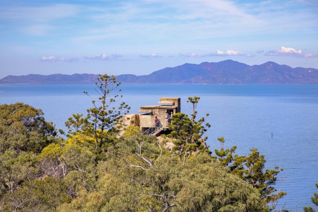 An old concrete bunker sits on a hill surrounded by trees, overlooking a calm blue sea with distant mountains under a partly cloudy sky.