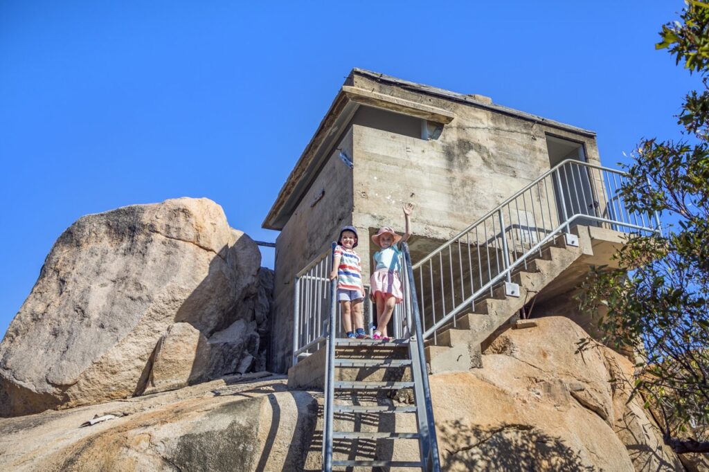 Two children stand on a metal staircase leading to a small stone structure built among large rocks. They are smiling and waving, with a clear blue sky in the background.