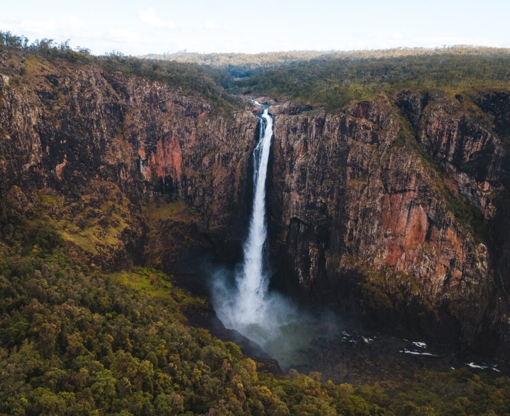 A tall waterfall cascades down steep cliffs into a lush green forested area, with a vast landscape in the background.