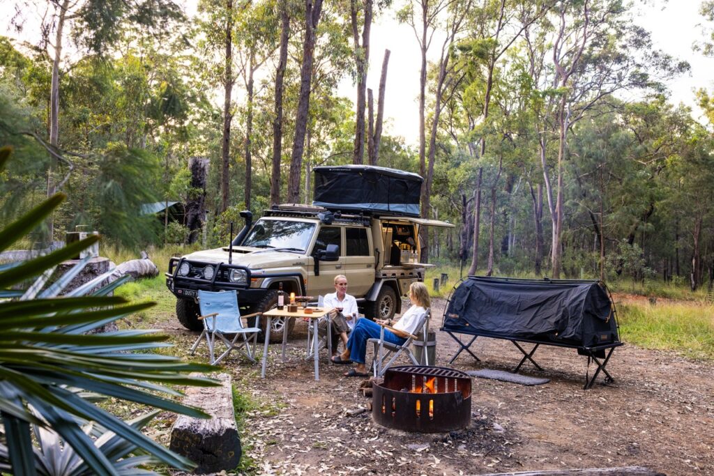 Two people sit at a camp table next to a rugged vehicle with a rooftop tent in a forested area. Camping equipment, including a small tent and a fire pit, are also present.