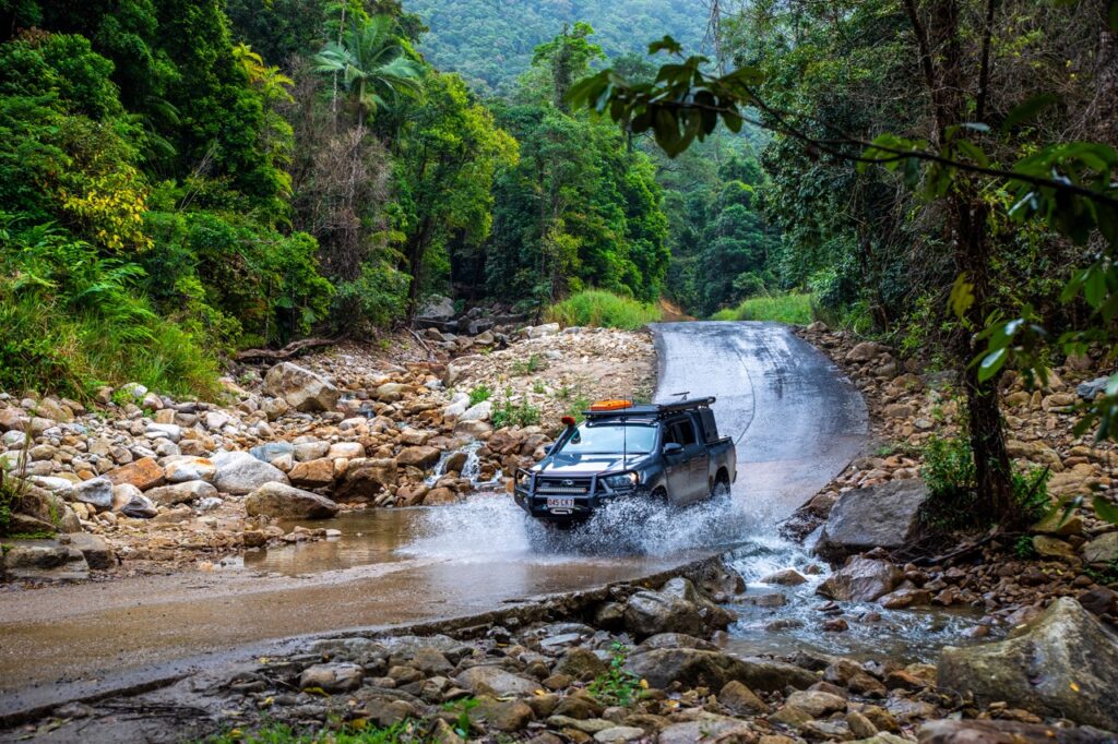 A truck with roof lights drives through a stream in a tropical forest, creating a splash, surrounded by green trees and rocky terrain with mountains in the background.