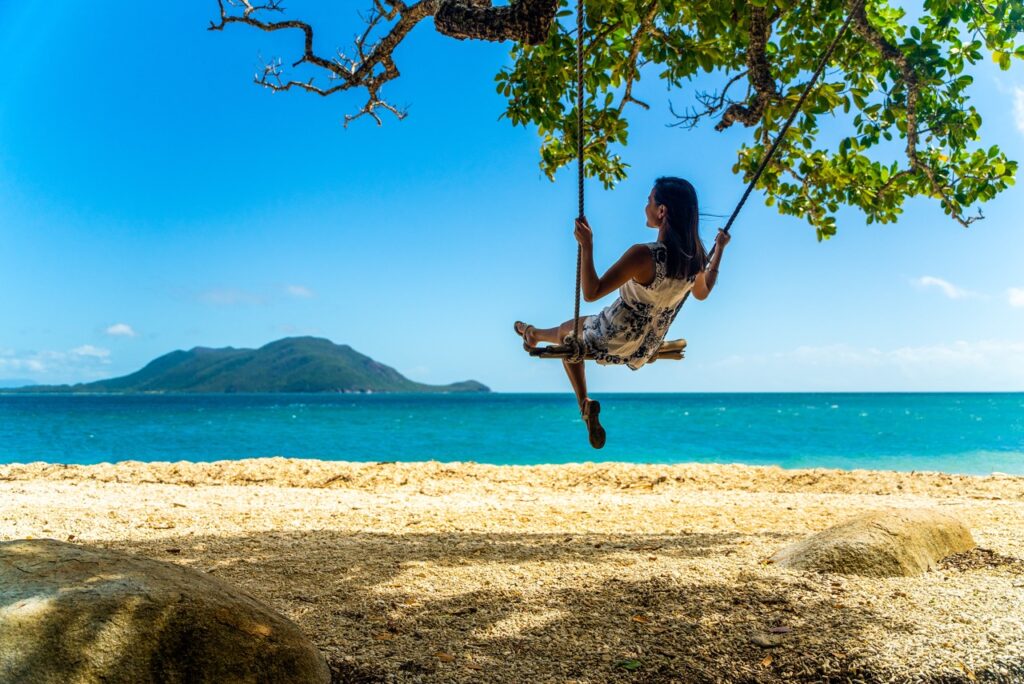 A person is sitting on a swing hanging from a tree by the beach, with an island and the ocean in the background under a clear blue sky.