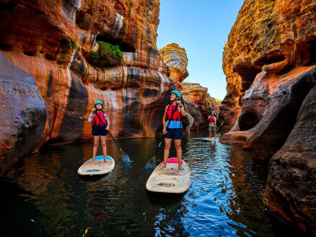 Three people paddleboarding through a narrow canyon with high rock walls. They are wearing life vests and helmets, navigating through calm water surrounded by rugged terrain.