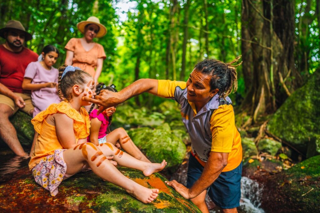 A group of people sits in the forest as an adult applies face paint to a child, with other children nearby.