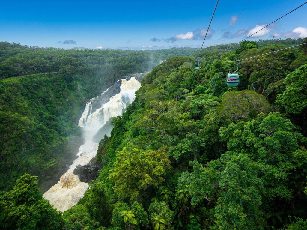 A cable car travels over a dense forested area near a powerful waterfall, with mist rising from the cascading water below.