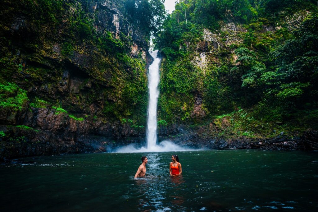 Two people stand in a natural pool at the base of a tall waterfall surrounded by lush greenery and rocky cliffs.